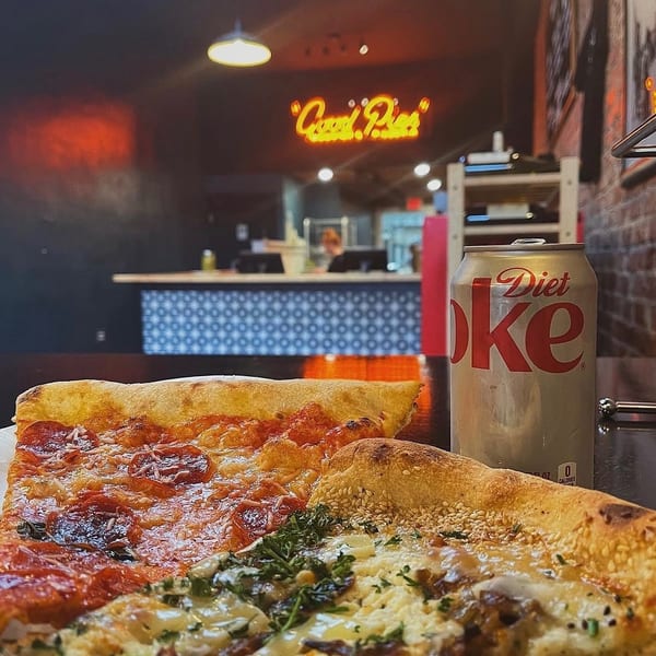 Two pizza slices and a Diet Coke can on a table with a neon sign in the background.