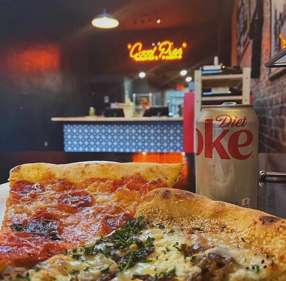 Two pizza slices and a Diet Coke can on a table with a neon sign in the background.