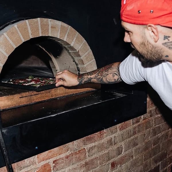 A chef in a red hat and white t-shirt placing a pizza into a brick oven.