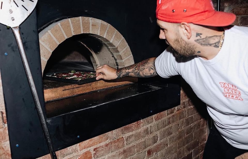 A chef in a red hat and white t-shirt placing a pizza into a brick oven.