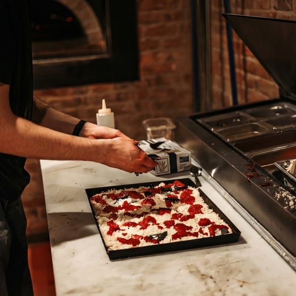 Chef preparing a square pizza with red sauce and white cheese on a marble counter.
