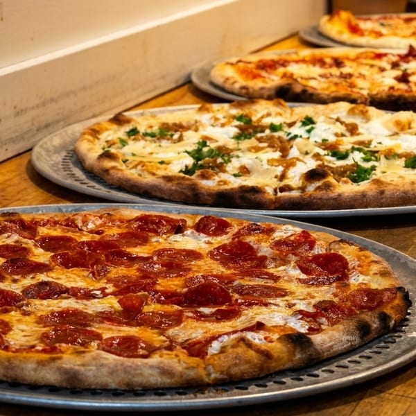 Four freshly baked pizzas, including pepperoni and white pizza, lined up on a rustic wooden counter.